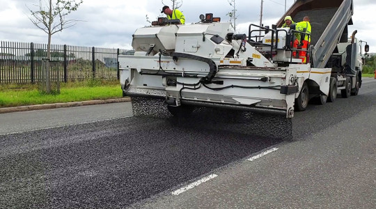A road receiving a stage of a surface dressing treatment, as large machinery lays chips on top of a layer of sticky bitumen