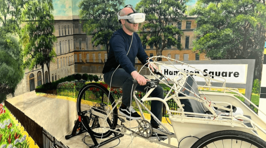 A person on a static bike with a VR headset on with the a staged background of Hamilton Square behind them.