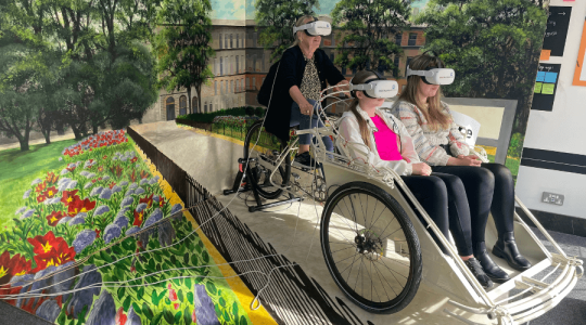 People using the Virtual Reality experience. One person is cycling on a static bike and two younger people are sitting in a set. All three people are wearing VR headsets with and behind them in a painted background of Hamilton Square. 