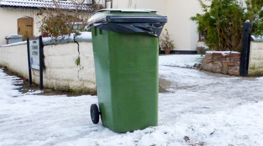 Green wheelie bin surrounded by snow 