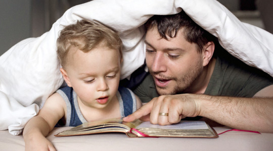 Dad reading book to son under bed covers