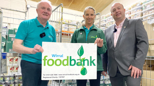 Photo of the foodbank manager, a foodbank volunteer and the Wirral Council Leader in Wirral foodbank holding a foodbank sign.