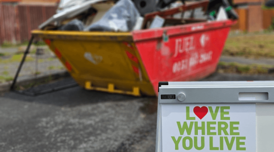A sign saying 'Love where you live' with a full skip behind it