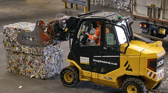 A yellow vehicle lifting a large bail of recycling in a recycling facility 