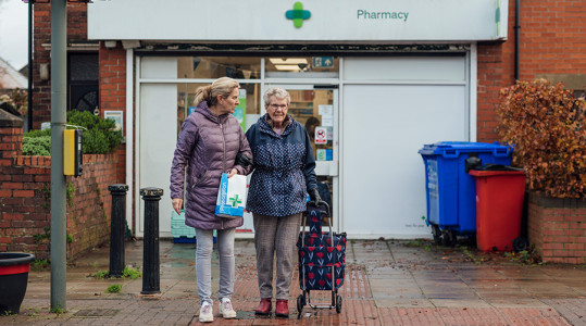 Image of a middle aged lady helping an older lady across a street outside a pharmacy