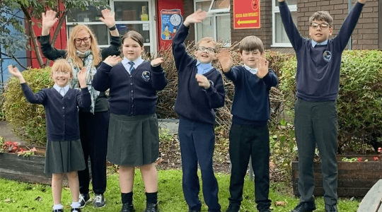 Five primary school pupils and their teacher with their arms in the air waving