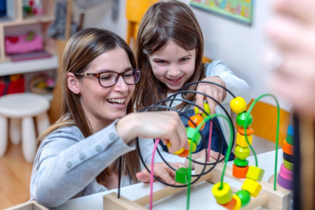 A woman with long brown hair in a grey jumper playing with a young girl with dark brown hair. They are both smiling.