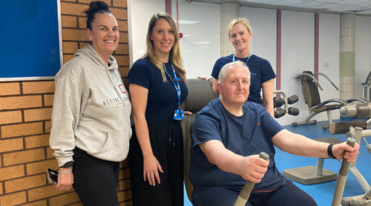 Barry sitting down using equipment with 3 ladies standing around him who have supported him through his recovery journey