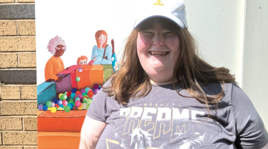 A young lady in a grey t shirt and white cap standing next to an art board. She is smiling.