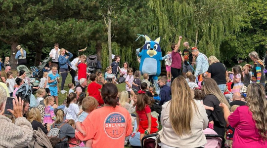Families gathered together in a park in front of a person dressed as the character Bluey