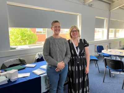 Two women from the PCPW steering group smiling in a bright room during an EBSA parental training session. They stand behind a table covered with a blue cloth displaying resources and guidance materials for parents. The setting includes other tables and chairs, indicating a welcoming group environment focused on supporting families with school attendance challenges.