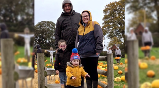 Four people standing on a wooden footbridge at a pumpkin patch. The group is dressed in warm, waterproof jackets and boots, with one child wearing a bright yellow coat and a colorful bobble hat. Pumpkins of various sizes are scattered across the grassy field and straw-covered paths in the background. Other visitors and wheelbarrows are visible among the pumpkins, with large trees and an overcast sky completing the autumn setting.