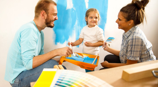Two adults and a child painting a wall bright blue together, using paint trays and brushes, with color swatches visible in the foreground