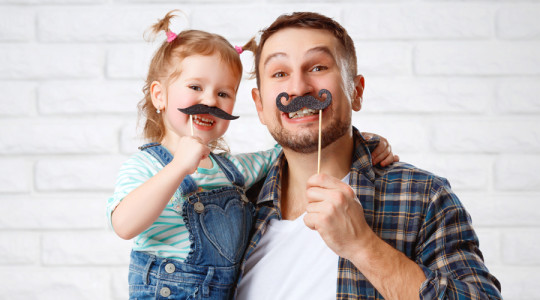 Smiling father and young daughter posing indoors against a white brick wall, each holding playful fake mustache props on sticks.