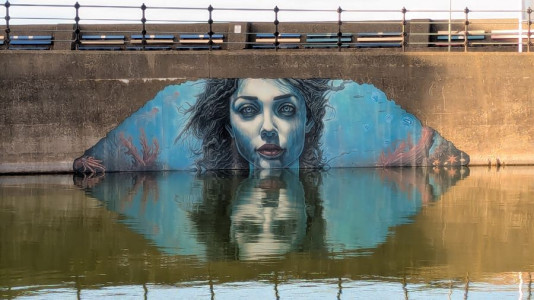 A mural painted on the side of a concrete bridge, depicting an underwater scene with blue tones, seaweed, coral, and marine life. The artwork is partially reflected in the calm water below, creating a mirrored effect. The bridge has a metal railing along the top and appears weathered, contrasting with the vibrant colors of the mural.