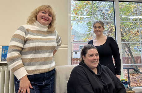 A group of adults seated around tables in a bright, modern classroom setting. They are attending a training session, with notebooks, pens, and printed materials on the tables. A presentation screen is visible in the background displaying course content. The attendees appear engaged, listening to a speaker who is out of frame.