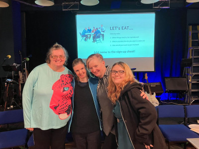 Four people standing together in a community venue with chairs in the background. A large screen behind them displays a slide titled “LET’s EAT…” with discussion points about tabletop talks and a sign-up sheet.