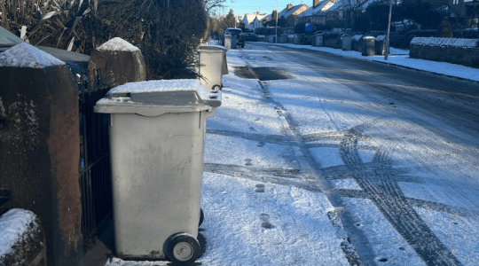 Side on view of a grey bin covered in snow with ice and snow on the pavement and road. 