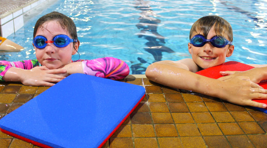 Girl and boy learning to swim arms resting on side of pool edge whilst in the water. A float aid is on ground in front of them