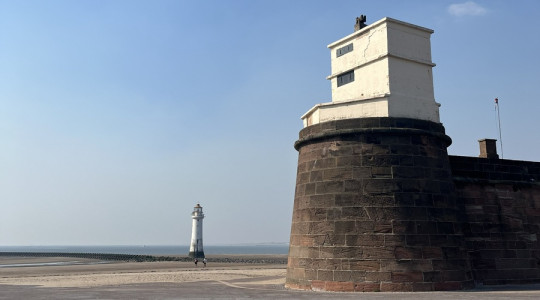 Fort Perch Rock on New Brighton seafront under a clear blue sky, with the stone fort in the foreground and the New Brighton Lighthouse visible in the distance across the beach and sea wall. A wide paved promenade stretches across the front of the image in bright daylight.