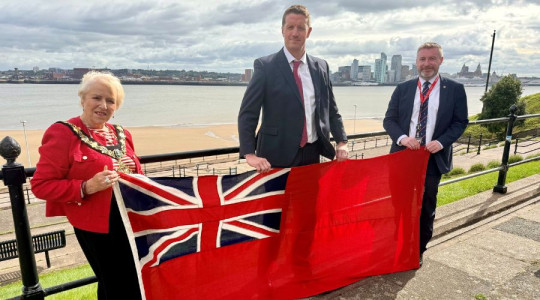 Photo of the Mayor and two representatives holding the Red Ensign with the River Mersey in the background