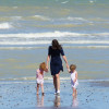A woman in a blue dress is walking along a beach holding the hands of two children