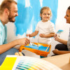Two adults and a child painting a wall bright blue together, using paint trays and brushes, with color swatches visible in the foreground