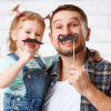 Smiling father and young daughter posing indoors against a white brick wall, each holding playful fake mustache props on sticks.