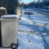 Side on view of a grey bin covered in snow with ice and snow on the pavement and road. 