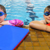 Girl and boy learning to swim arms resting on side of pool edge whilst in the water. A float aid is on ground in front of them