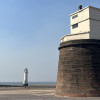 Fort Perch Rock on New Brighton seafront under a clear blue sky, with the stone fort in the foreground and the New Brighton Lighthouse visible in the distance across the beach and sea wall. A wide paved promenade stretches across the front of the image in bright daylight.