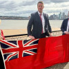 Photo of the Mayor and two representatives holding the Red Ensign with the River Mersey in the background