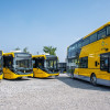 Fleet of Wirral's new yellow buses parked outdoors, including a double-decker and several single-decker vehicles, pictured in a depot under a clear blue sky.