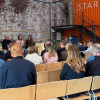 Audience members seated in rows of chairs listening to a panel discussion inside the Start-Yard venue. Speakers sit at the front against an exposed brick wall, with an orange container-style structure displaying the text “START-YARD” and a metal staircase in the background.