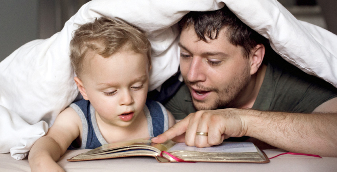 Dad reading book to son under bed covers