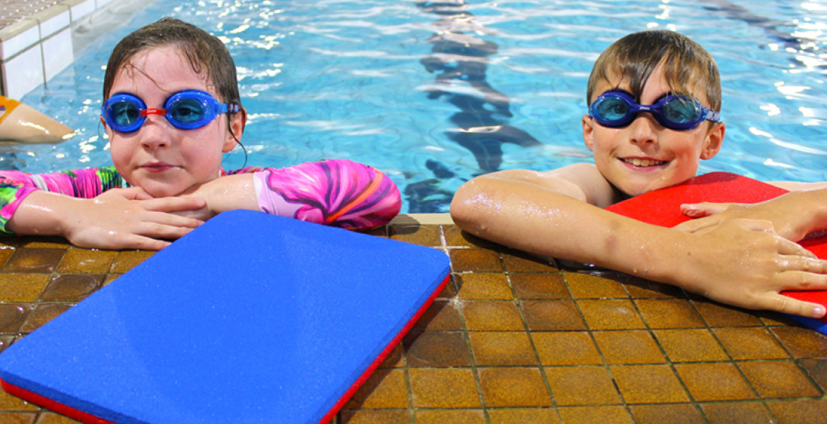 Girl and boy learning to swim arms resting on side of pool edge whilst in the water. A float aid is on ground in front of them