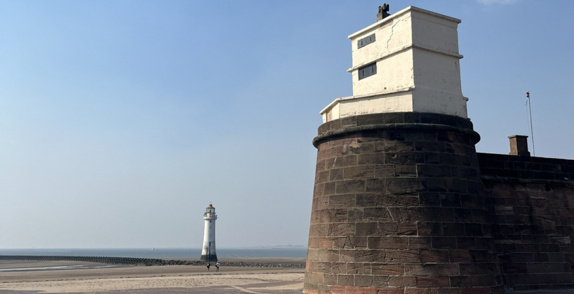 Fort Perch Rock on New Brighton seafront under a clear blue sky, with the stone fort in the foreground and the New Brighton Lighthouse visible in the distance across the beach and sea wall. A wide paved promenade stretches across the front of the image in bright daylight.
