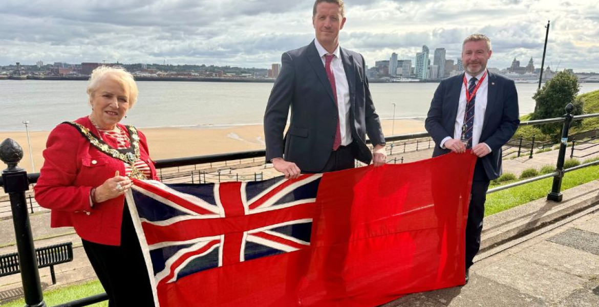 Photo of the Mayor and two representatives holding the Red Ensign with the River Mersey in the background