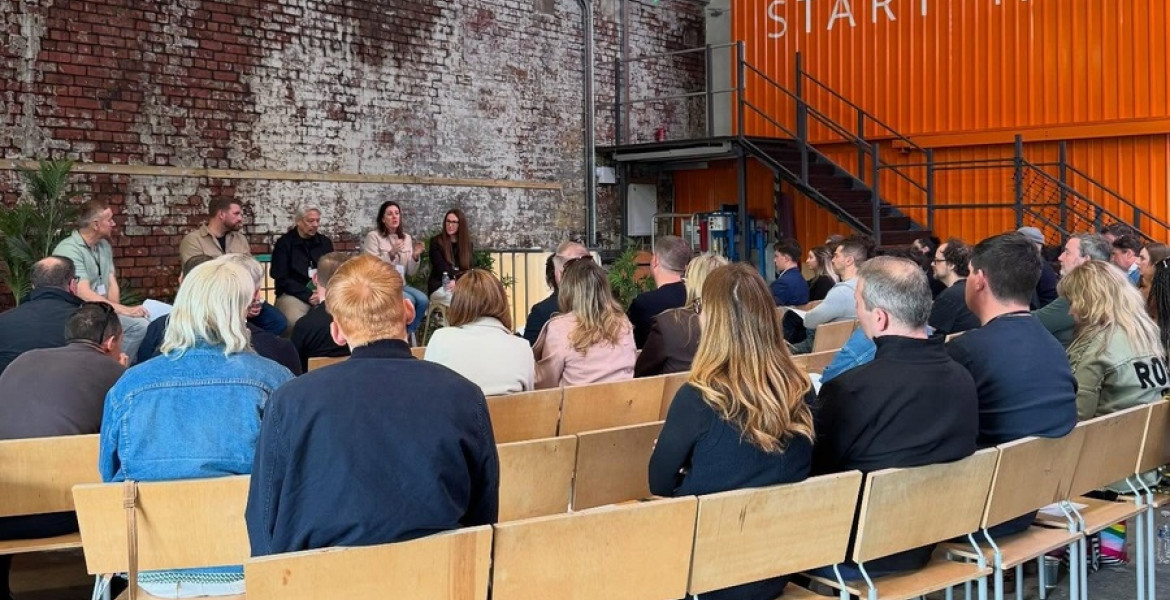 Audience members seated in rows of chairs listening to a panel discussion inside the Start-Yard venue. Speakers sit at the front against an exposed brick wall, with an orange container-style structure displaying the text “START-YARD” and a metal staircase in the background.
